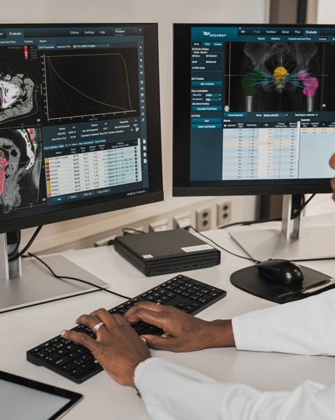 A radiologist sitting at a desk, looking at two computer screens showing scan software